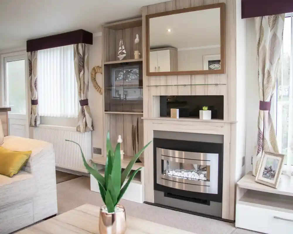 A cozy living room with a light color scheme features a modern fireplace with a mirror above it. There are built-in shelves with decorative items and a potted plant on the coffee table. Natural light streams in through the window, which has white blinds. A comfortable sofa adds to the inviting atmosphere.