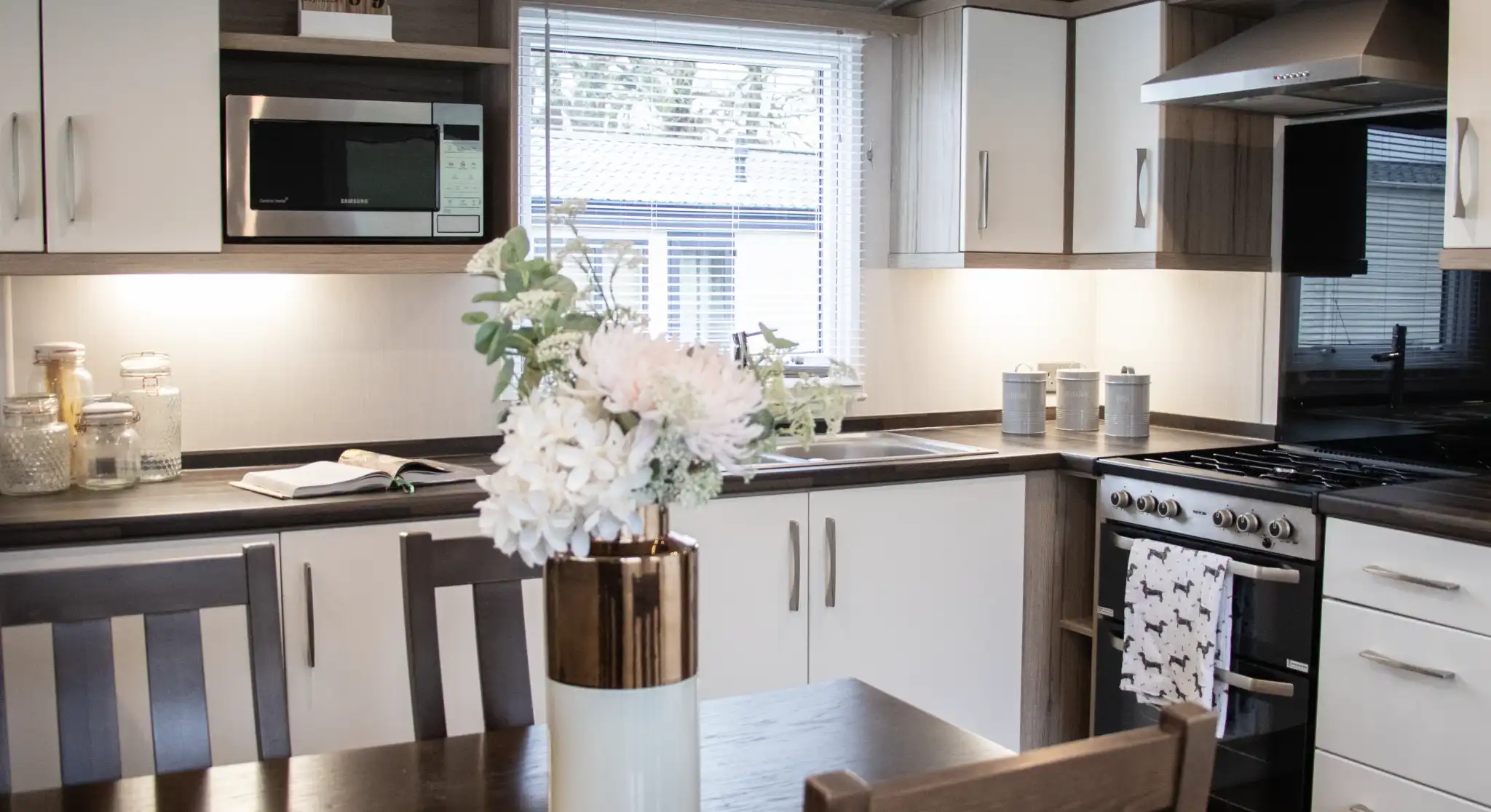 A modern kitchen featuring sleek cabinetry in white and dark wood, equipped with a microwave, oven, and a sink. A dining table with wooden chairs is in the foreground, adorned with a vase of flowers. Natural light streams in through a window.