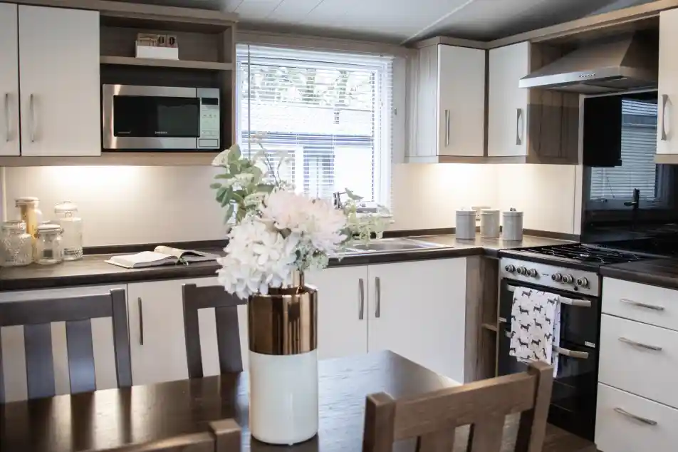 A modern kitchen featuring sleek cabinetry in white and dark wood, equipped with a microwave, oven, and a sink. A dining table with wooden chairs is in the foreground, adorned with a vase of flowers. Natural light streams in through a window.