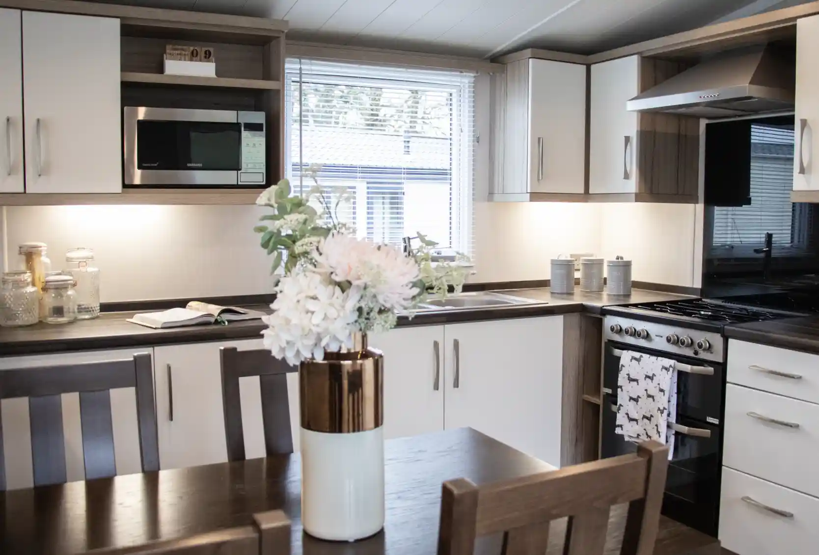 A modern kitchen featuring sleek cabinetry in white and dark wood, equipped with a microwave, oven, and a sink. A dining table with wooden chairs is in the foreground, adorned with a vase of flowers. Natural light streams in through a window.