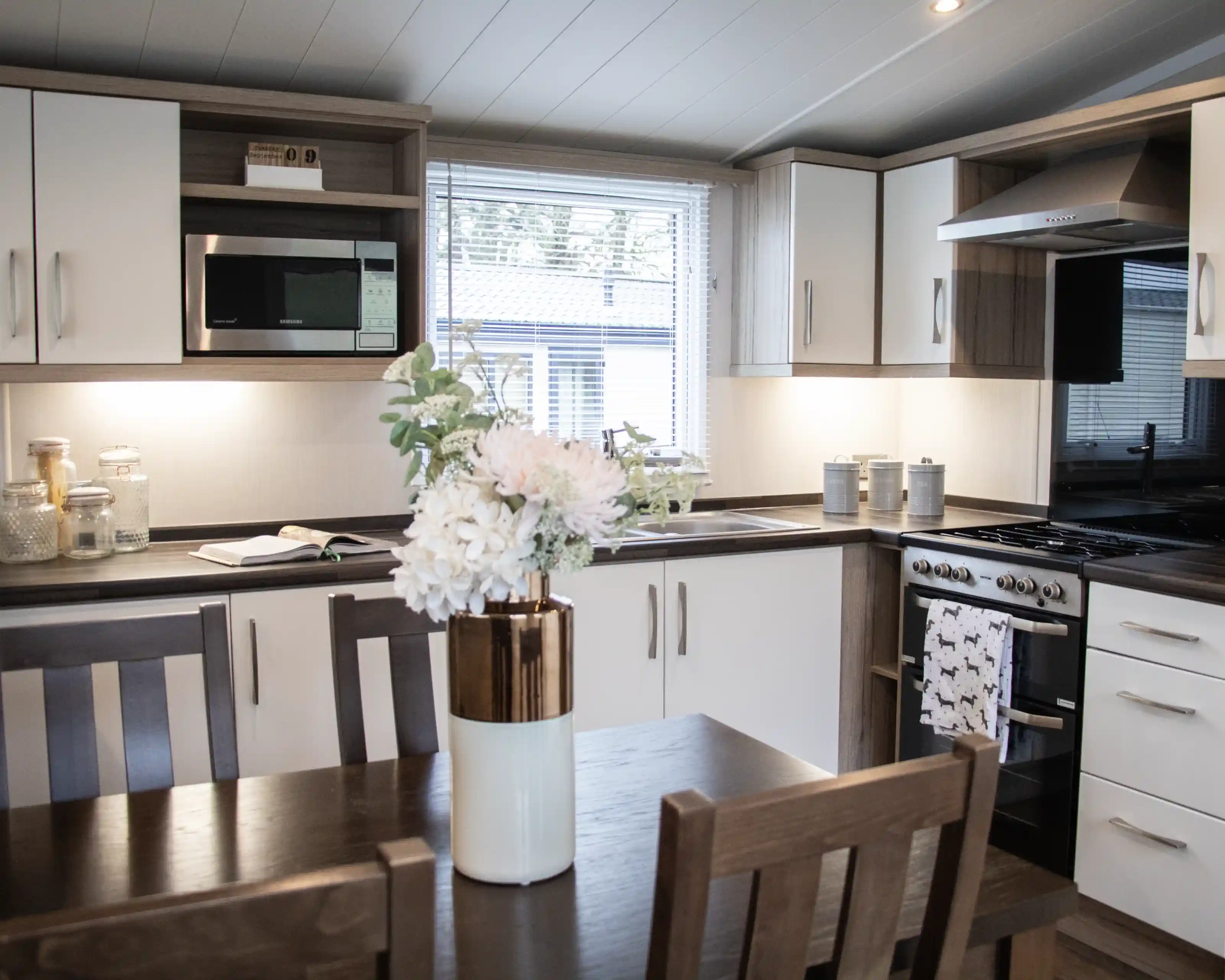 A modern kitchen featuring sleek cabinetry in white and dark wood, equipped with a microwave, oven, and a sink. A dining table with wooden chairs is in the foreground, adorned with a vase of flowers. Natural light streams in through a window.