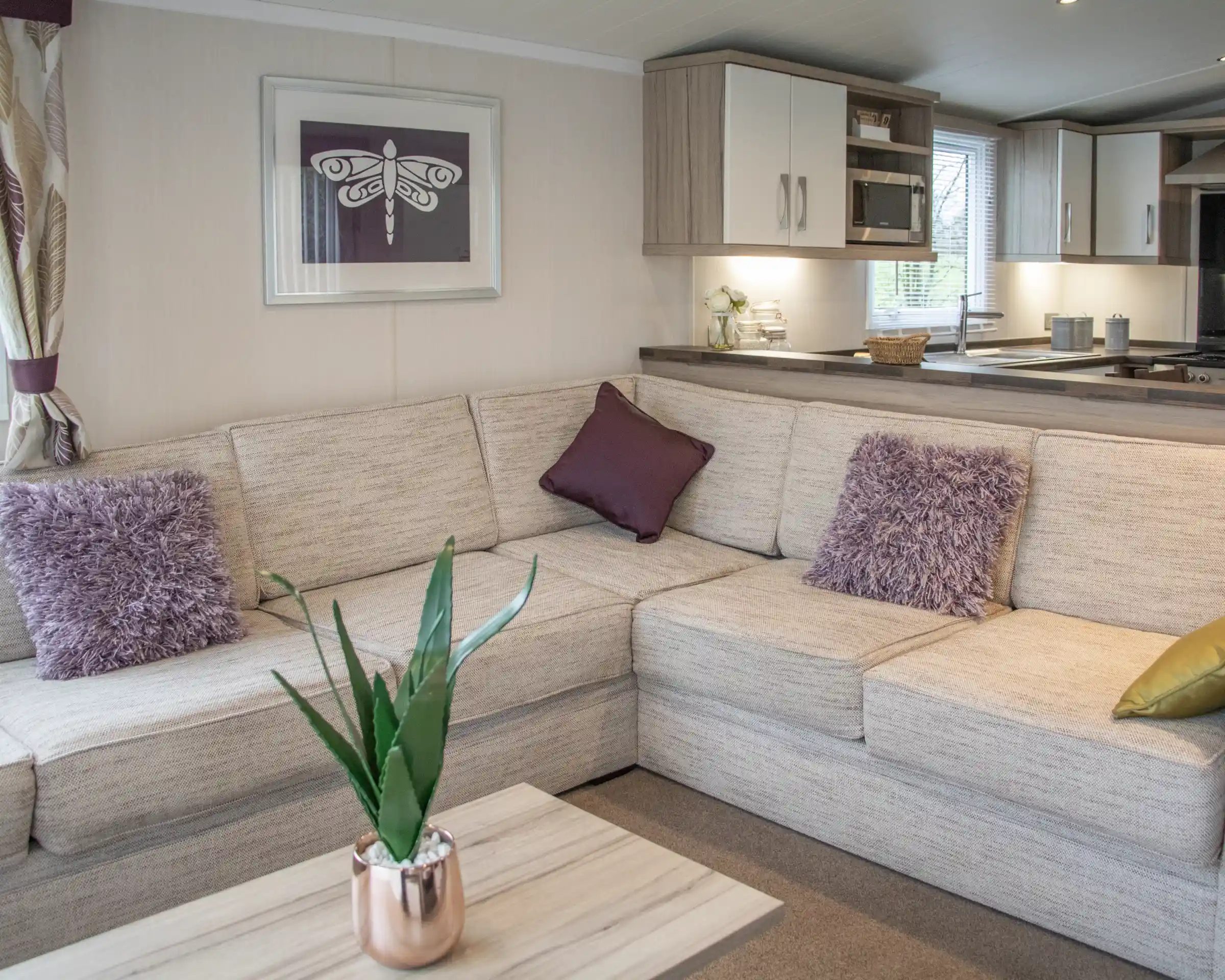 A cozy living space featuring a light-colored sectional sofa adorned with purple throw pillows and a decorative plant. A wooden coffee table sits in front, and a modern kitchen area is visible in the background.