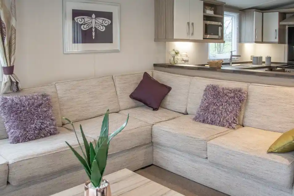 A cozy living space featuring a light-colored sectional sofa adorned with purple throw pillows and a decorative plant. A wooden coffee table sits in front, and a modern kitchen area is visible in the background.