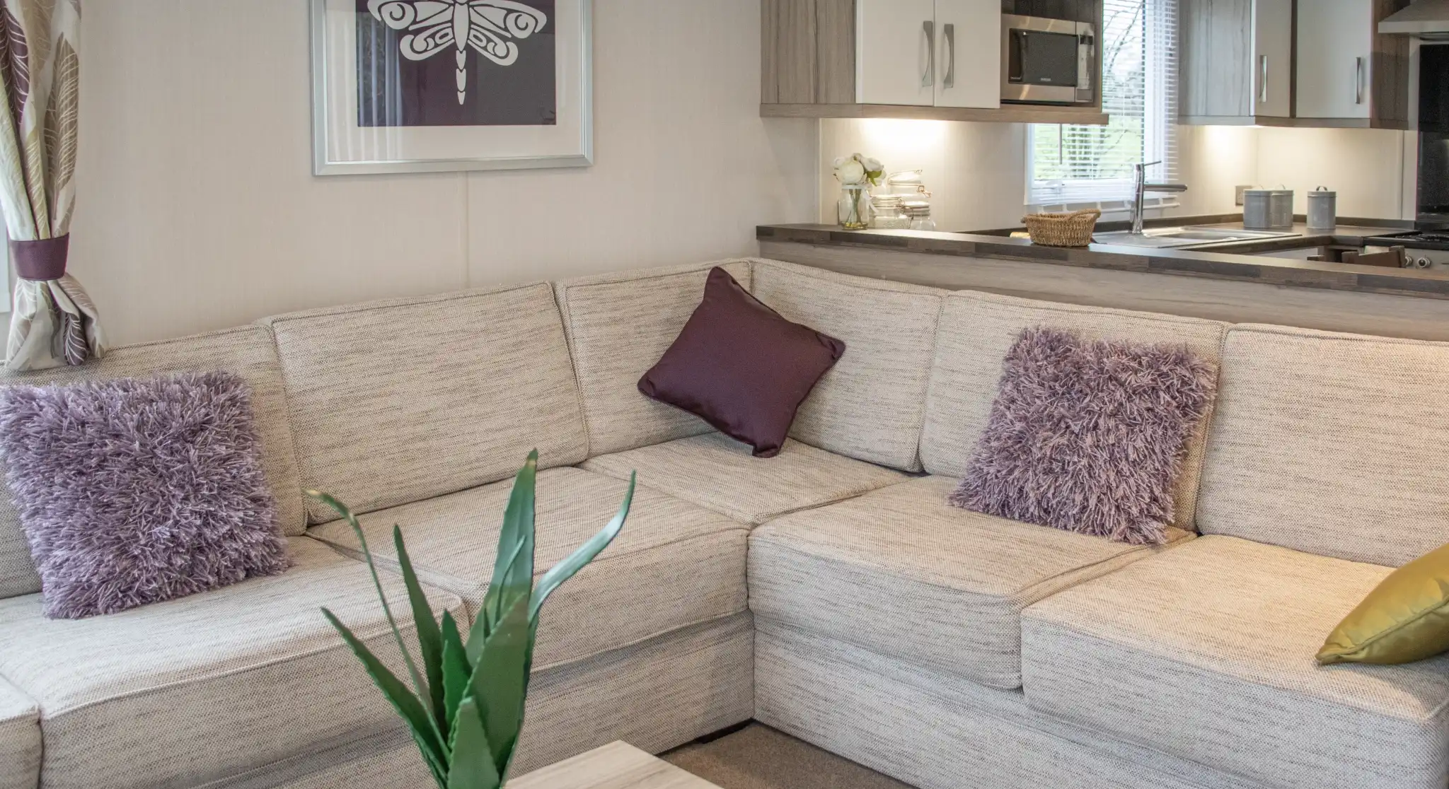 A cozy living space featuring a light-colored sectional sofa adorned with purple throw pillows and a decorative plant. A wooden coffee table sits in front, and a modern kitchen area is visible in the background.