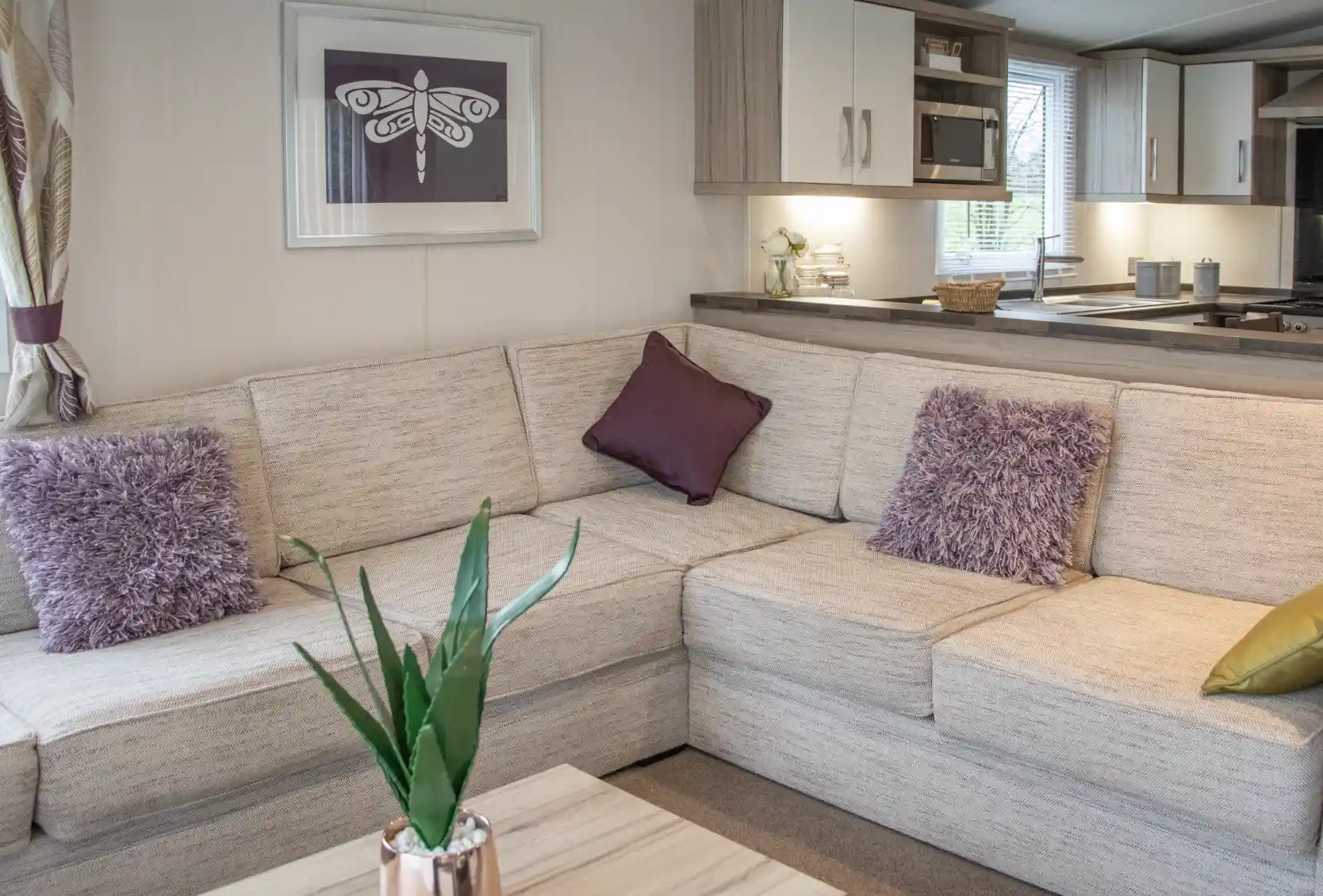 A cozy living space featuring a light-colored sectional sofa adorned with purple throw pillows and a decorative plant. A wooden coffee table sits in front, and a modern kitchen area is visible in the background.
