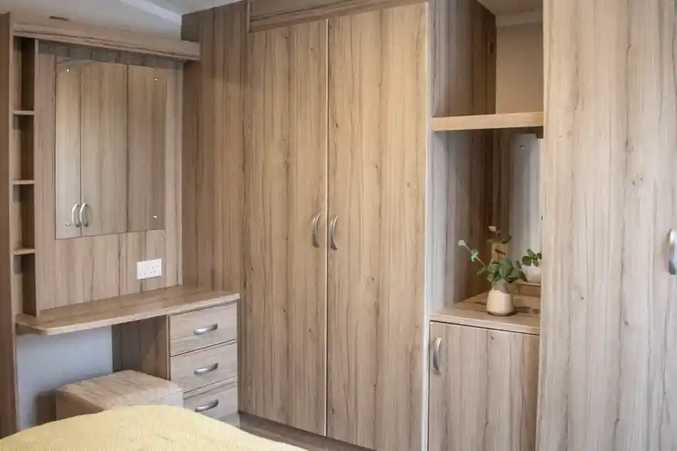 A cozy bedroom featuring light wood cabinets, a mirror, and a small desk with drawers. A plant sits on the surface near the mirror. The bedding is a soft yellow.