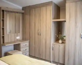 A cozy bedroom featuring light wood cabinets, a mirror, and a small desk with drawers. A plant sits on the surface near the mirror. The bedding is a soft yellow.