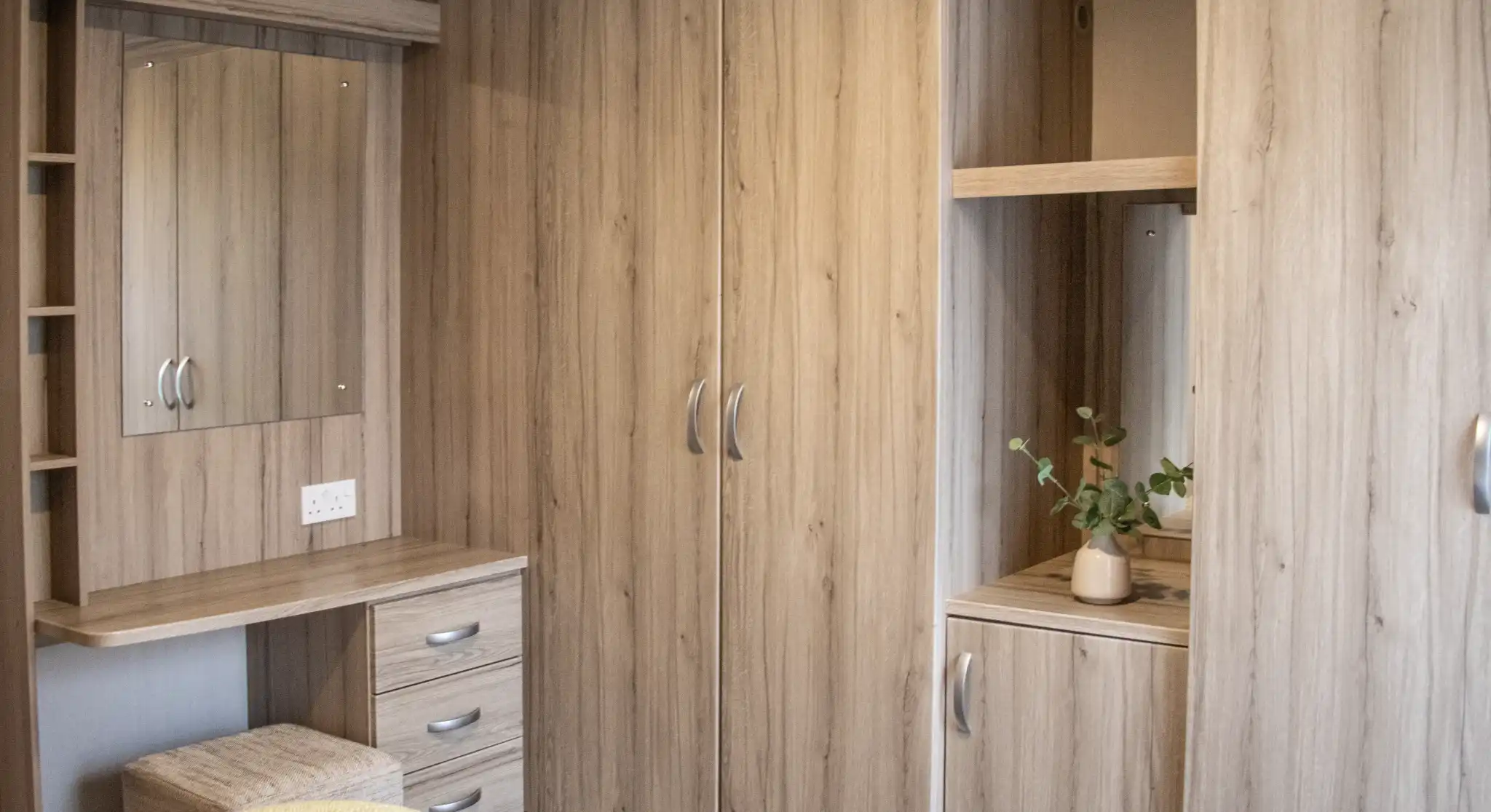 A cozy bedroom featuring light wood cabinets, a mirror, and a small desk with drawers. A plant sits on the surface near the mirror. The bedding is a soft yellow.