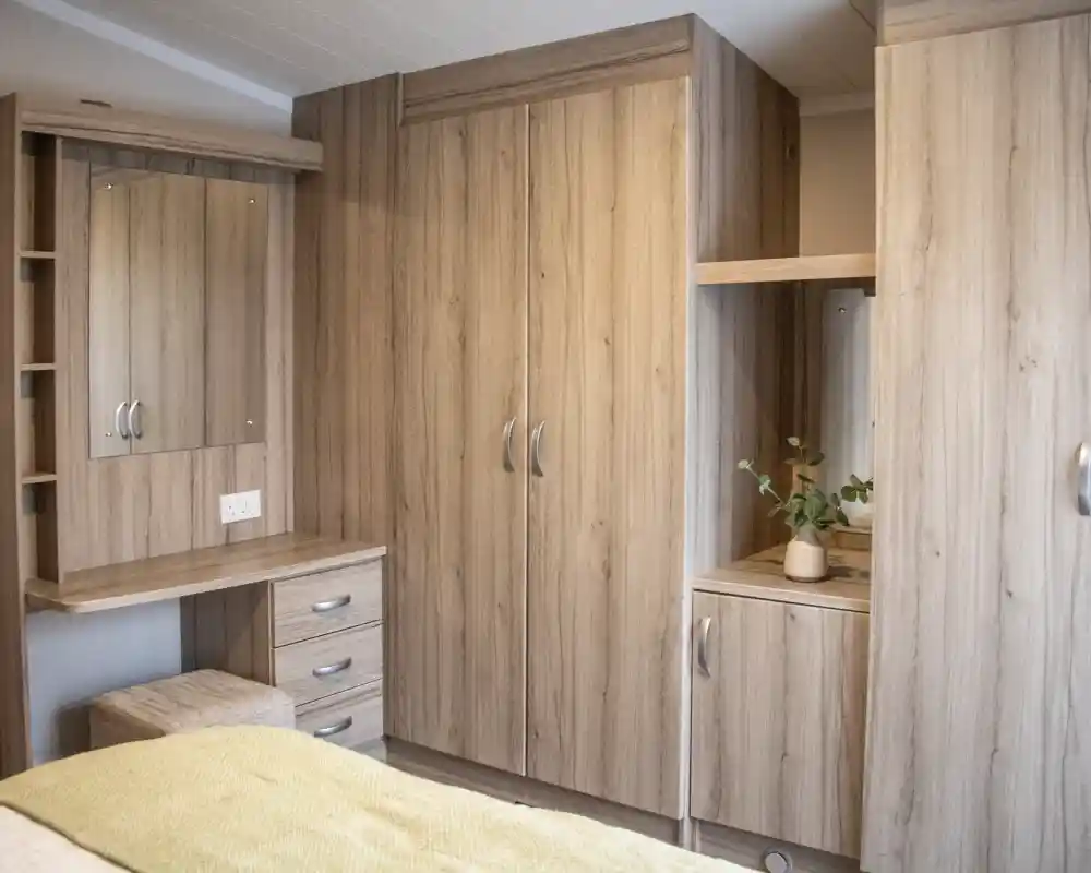A cozy bedroom featuring light wood cabinets, a mirror, and a small desk with drawers. A plant sits on the surface near the mirror. The bedding is a soft yellow.