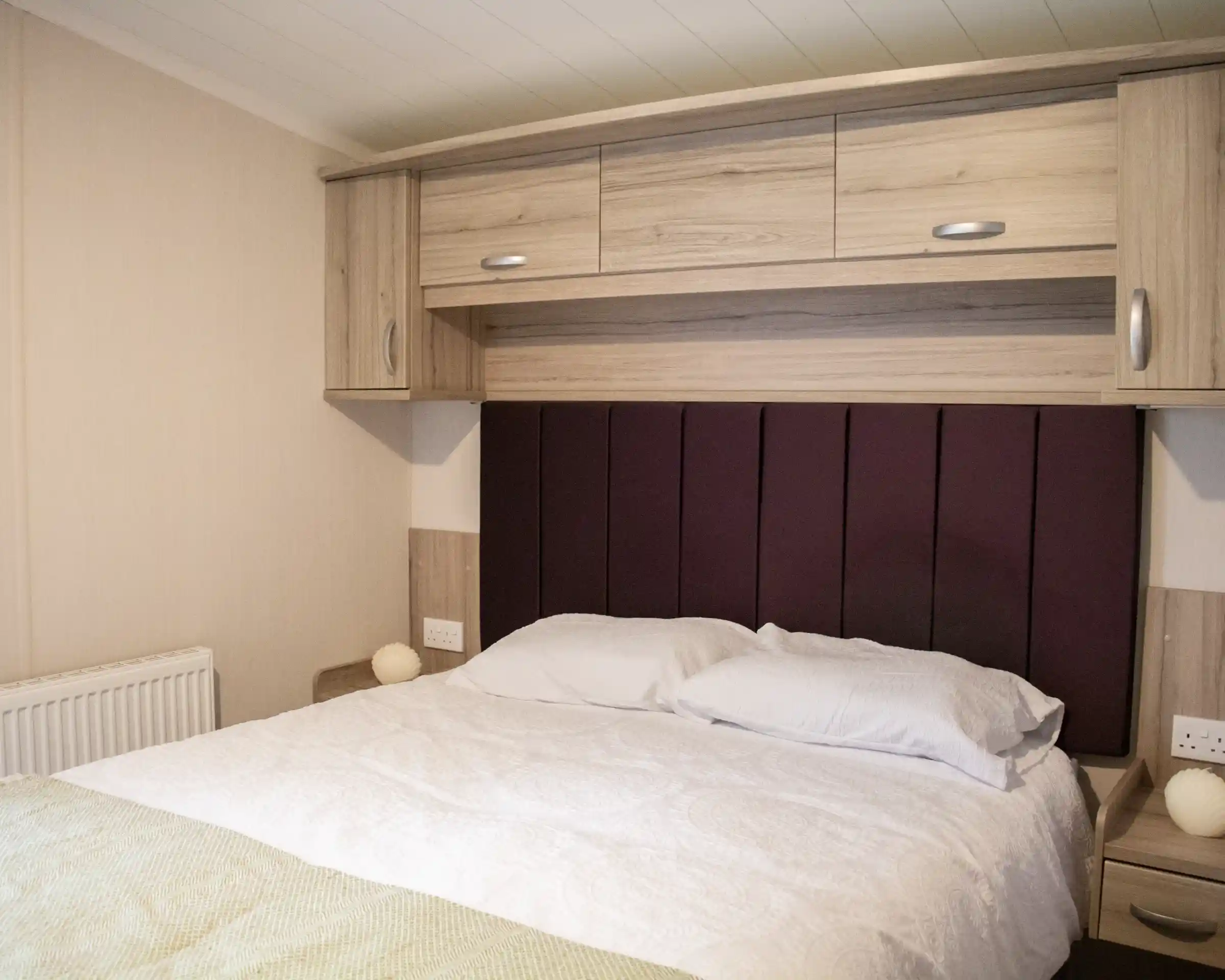 A cozy bedroom featuring a bed with white bedding and a purple upholstered headboard. Wooden cabinets and bedside tables are on either side, and there is a radiator and soft lighting in the room.