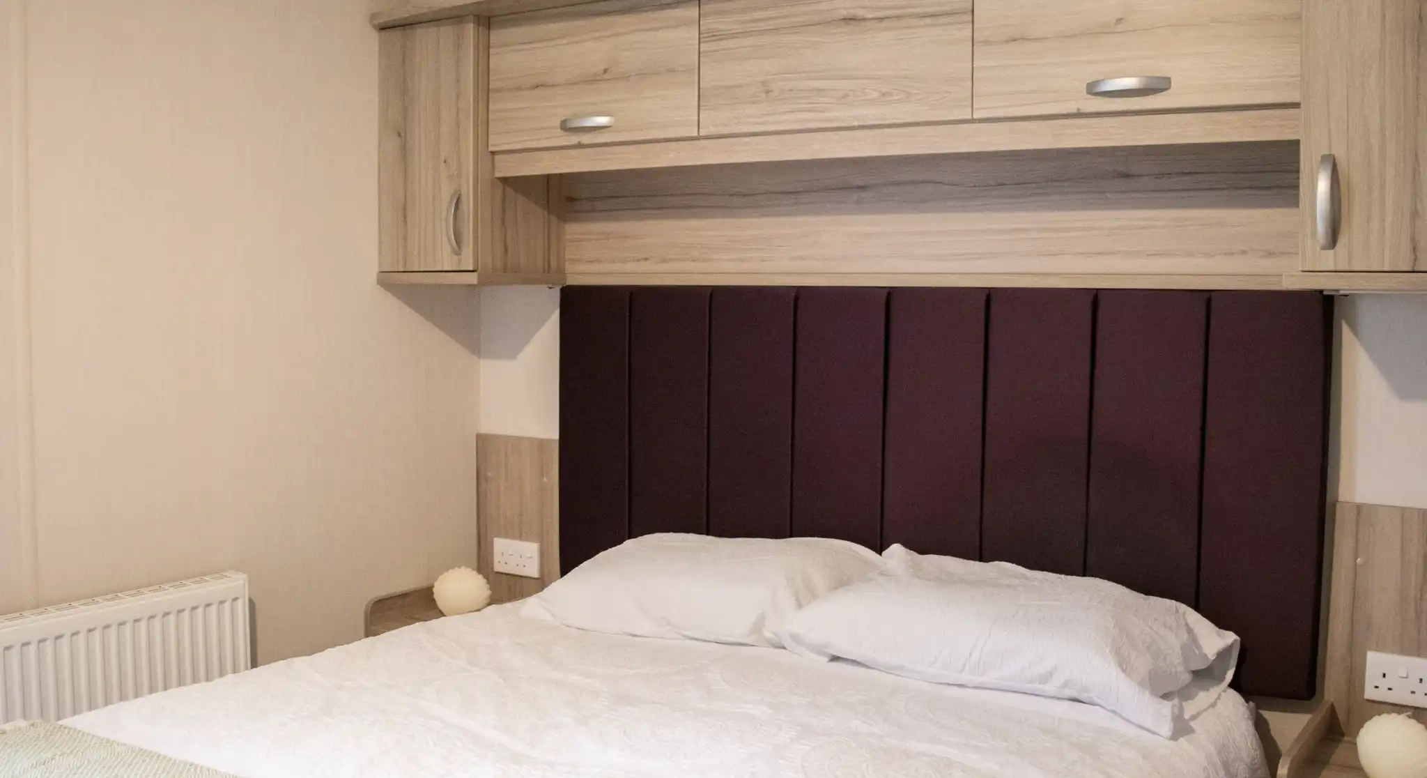 A cozy bedroom featuring a bed with white bedding and a purple upholstered headboard. Wooden cabinets and bedside tables are on either side, and there is a radiator and soft lighting in the room.