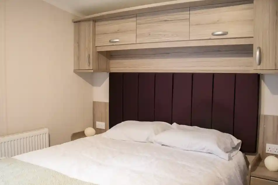 A cozy bedroom featuring a bed with white bedding and a purple upholstered headboard. Wooden cabinets and bedside tables are on either side, and there is a radiator and soft lighting in the room.