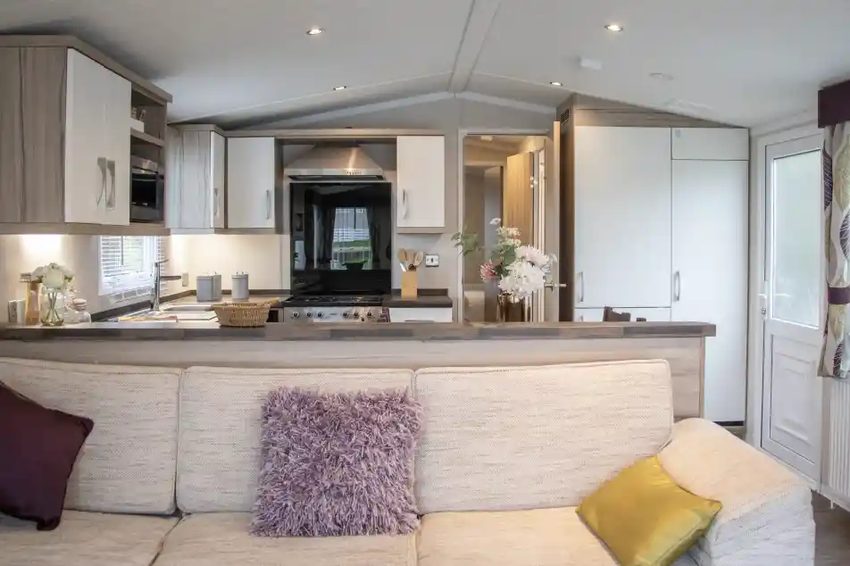 A cozy living room with a beige sofa adorned with purple and yellow cushions. In the background, a modern kitchen features wooden cabinets, a black stove, and a dining area. Natural light pours in through the large windows.
