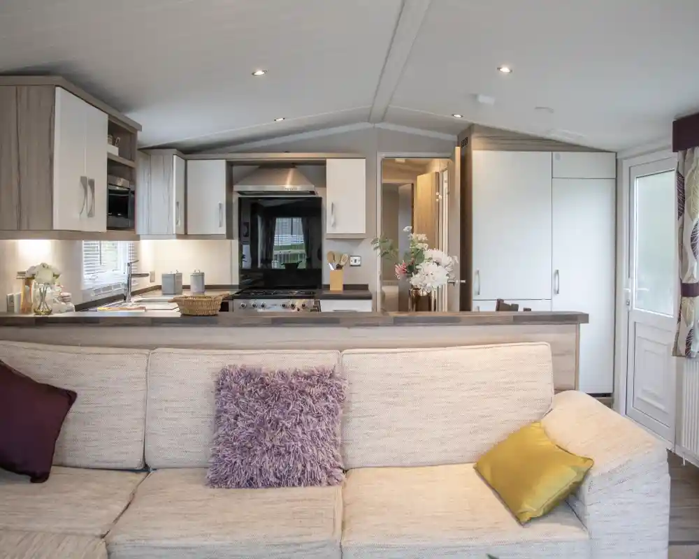 A cozy living room with a beige sofa adorned with purple and yellow cushions. In the background, a modern kitchen features wooden cabinets, a black stove, and a dining area. Natural light pours in through the large windows.