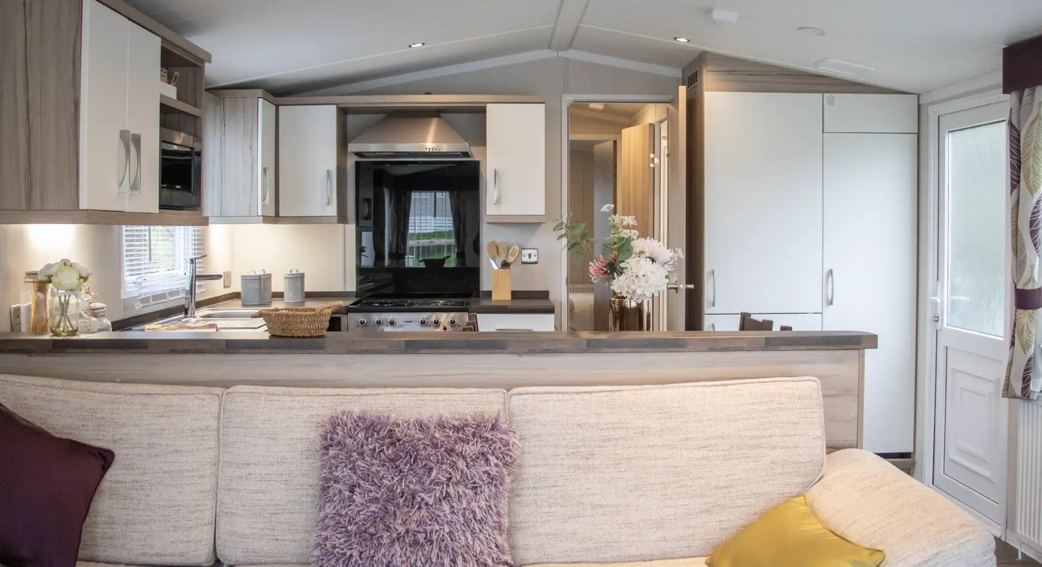 A cozy living room with a beige sofa adorned with purple and yellow cushions. In the background, a modern kitchen features wooden cabinets, a black stove, and a dining area. Natural light pours in through the large windows.