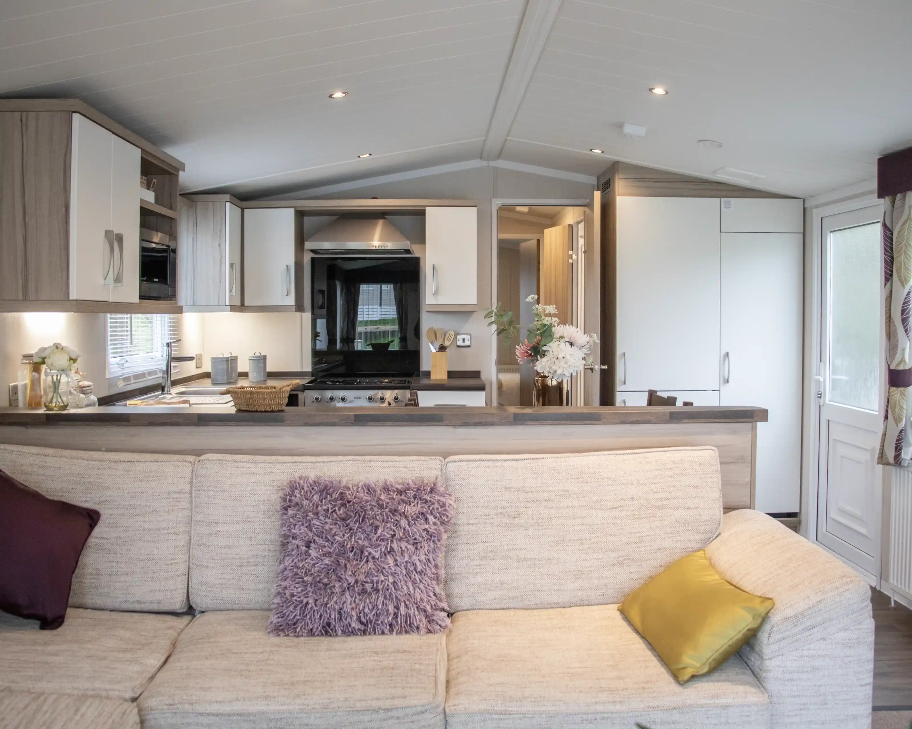 A cozy living room with a beige sofa adorned with purple and yellow cushions. In the background, a modern kitchen features wooden cabinets, a black stove, and a dining area. Natural light pours in through the large windows.