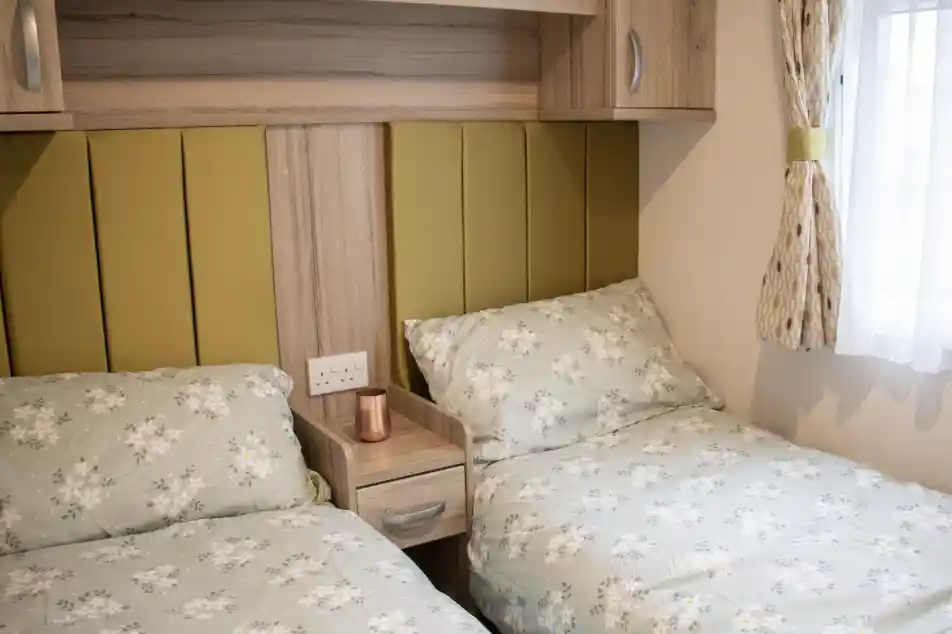 A cozy bedroom with two twin beds featuring floral patterned bedding. A wooden nightstand with a copper mug sits between the beds. Soft green accents on the headboards and warm lighting create a welcoming atmosphere.