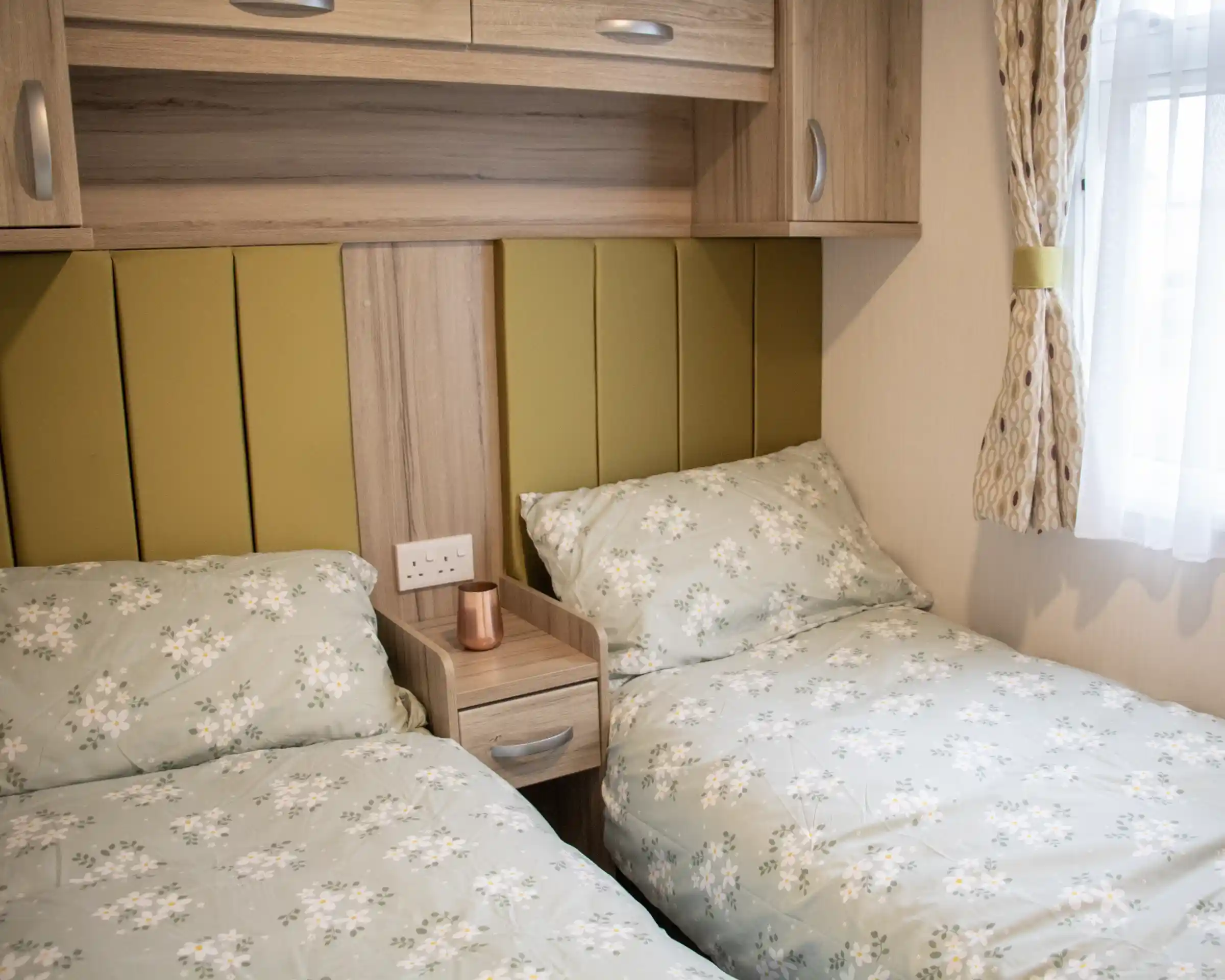 A cozy bedroom with two twin beds featuring floral patterned bedding. A wooden nightstand with a copper mug sits between the beds. Soft green accents on the headboards and warm lighting create a welcoming atmosphere.