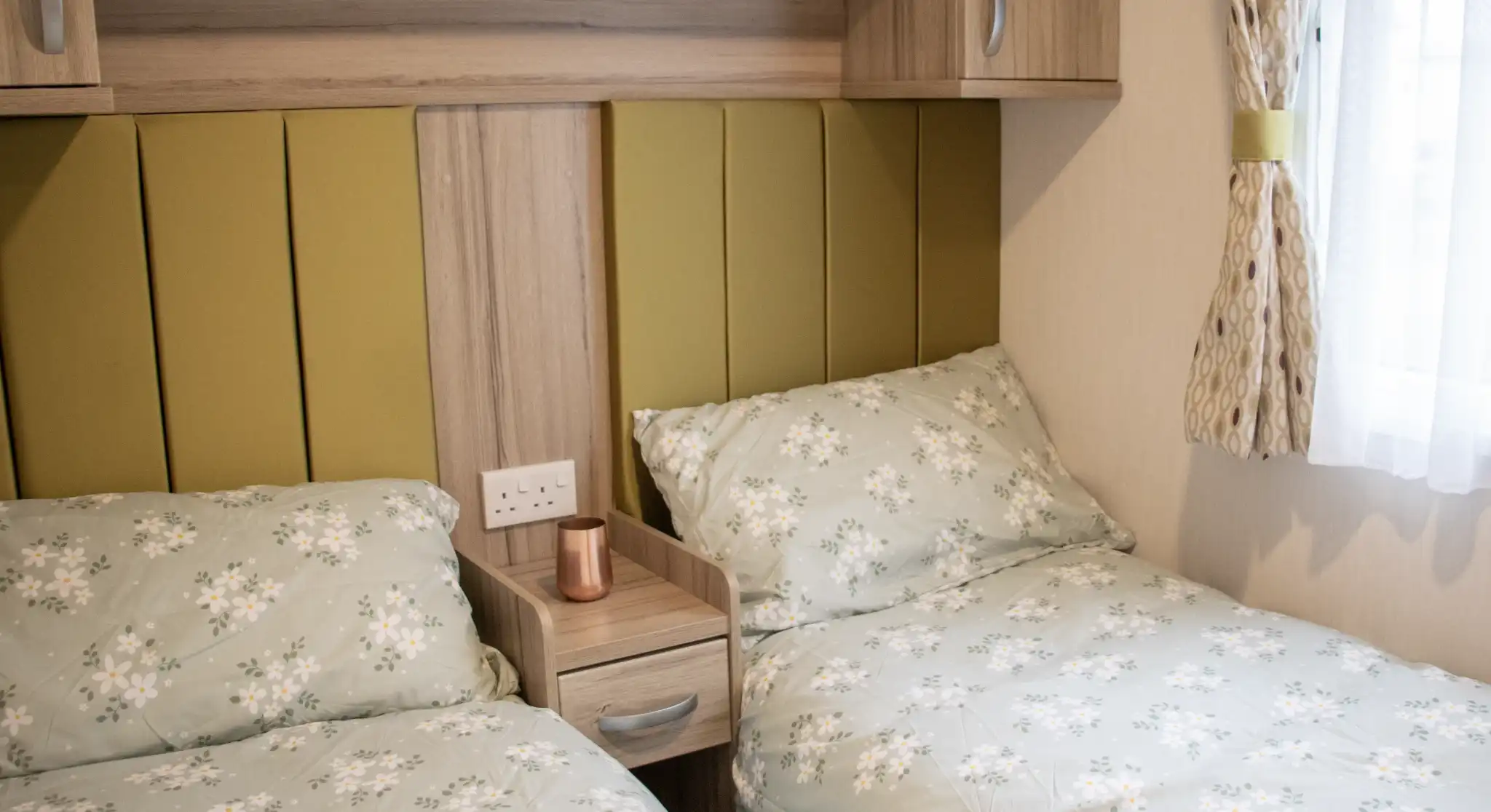 A cozy bedroom with two twin beds featuring floral patterned bedding. A wooden nightstand with a copper mug sits between the beds. Soft green accents on the headboards and warm lighting create a welcoming atmosphere.