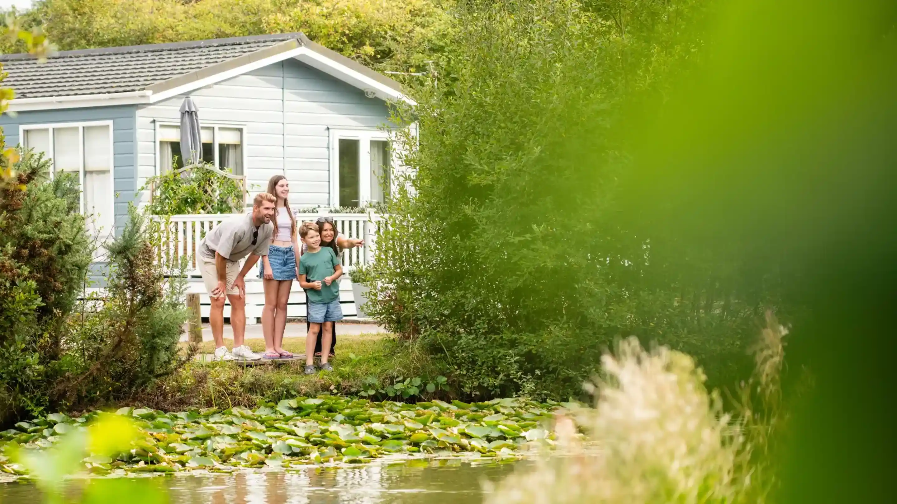 A family stands by a pond covered with lily pads, smiling and looking at something in the water. In the background, a light blue cabin is partially visible surrounded by greenery.
