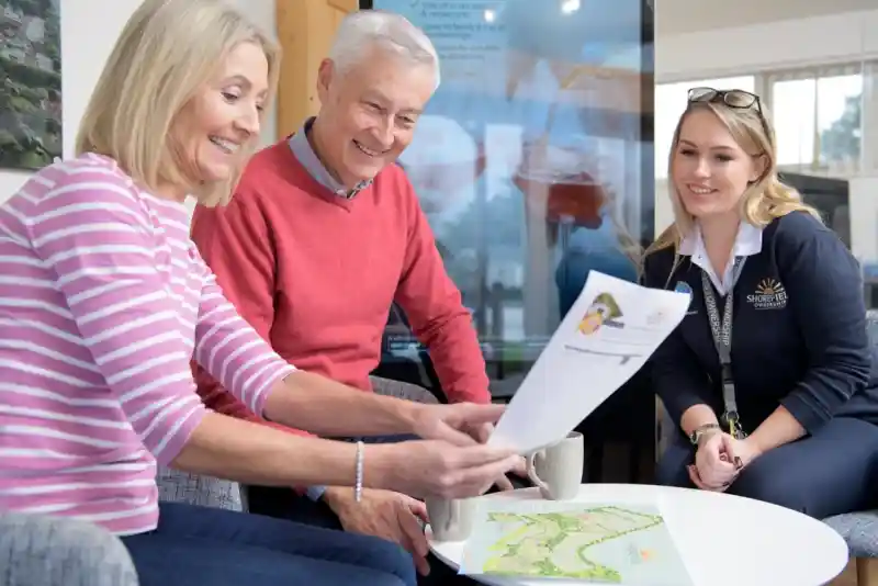 Two older adults and a young woman are sitting together, smiling and reviewing a document at a table. They are in a modern, bright setting with a screen displaying images in the background. Each person has a cup of tea or coffee.