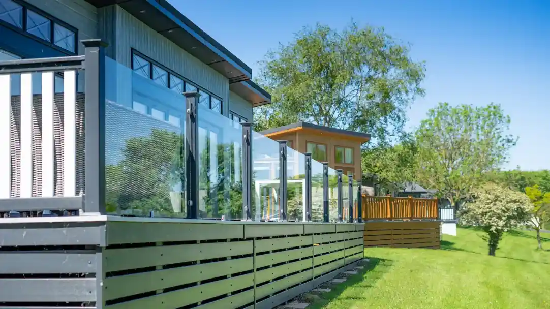 A modern house features a sleek deck with glass railings overlooking a green lawn. Various trees and another wooden structure are visible in the background under a clear blue sky.