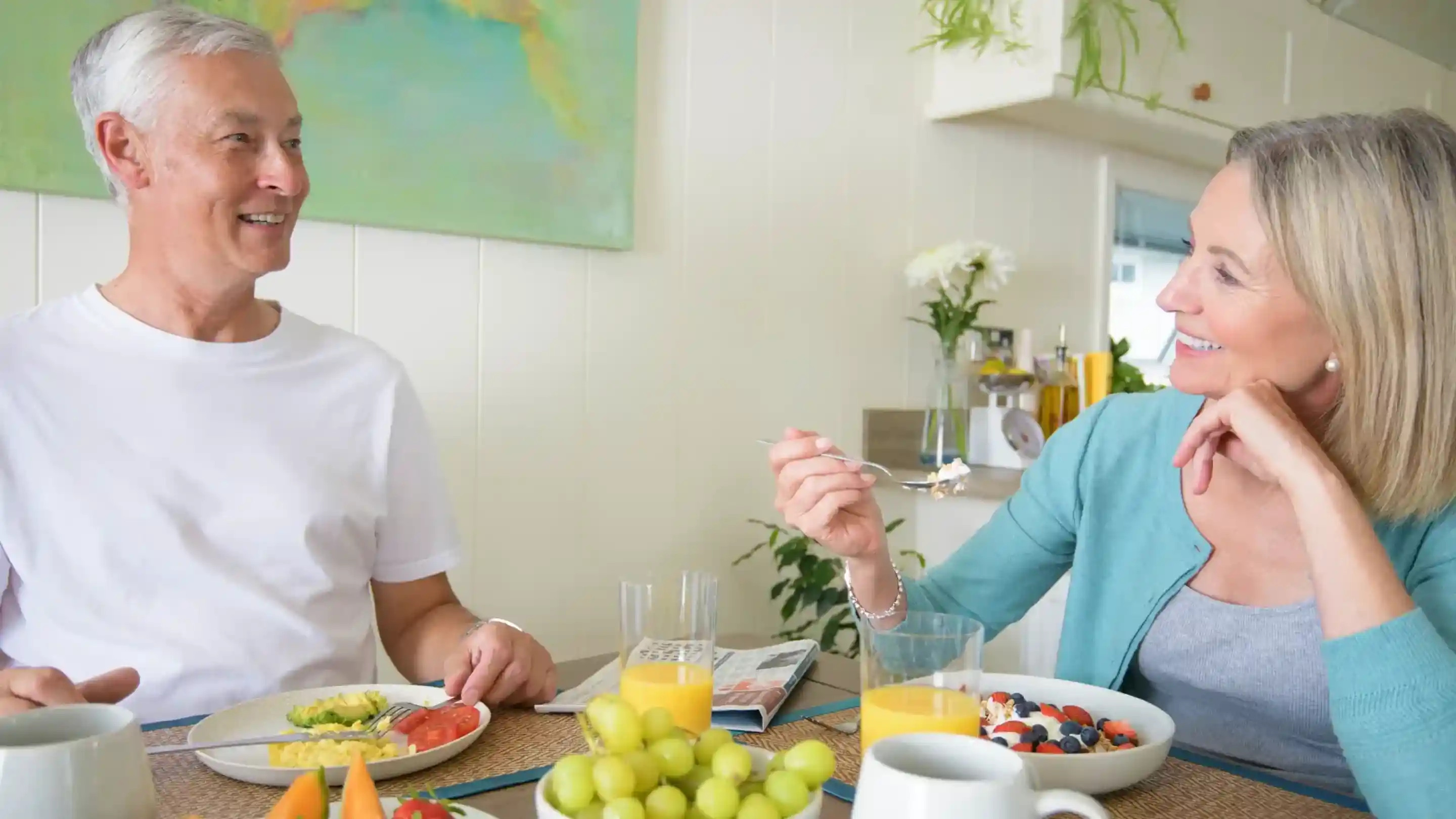 A man and woman enjoy breakfast together at a kitchen table, smiling and engaging in conversation. The table is adorned with colorful plates of food, including fruit and eggs, along with glasses of juice. A vibrant painting hangs in the background.