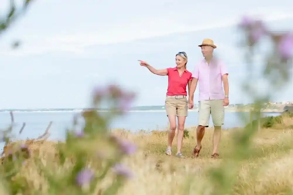 A couple walks hand in hand along a scenic coastal path, with the woman pointing out something in the distance. She wears a pink polo shirt and shorts, while the man is in a light pink shirt and beige shorts. Wildflowers frame the scene, adding a natural touch to the landscape.