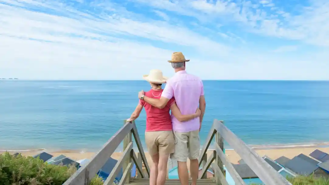 A couple stands on a wooden staircase overlooking the ocean, wearing summer attire and straw hats. They are embracing, enjoying a scenic view of the calm blue waters and a sandy beach below, with a clear sky and light clouds.