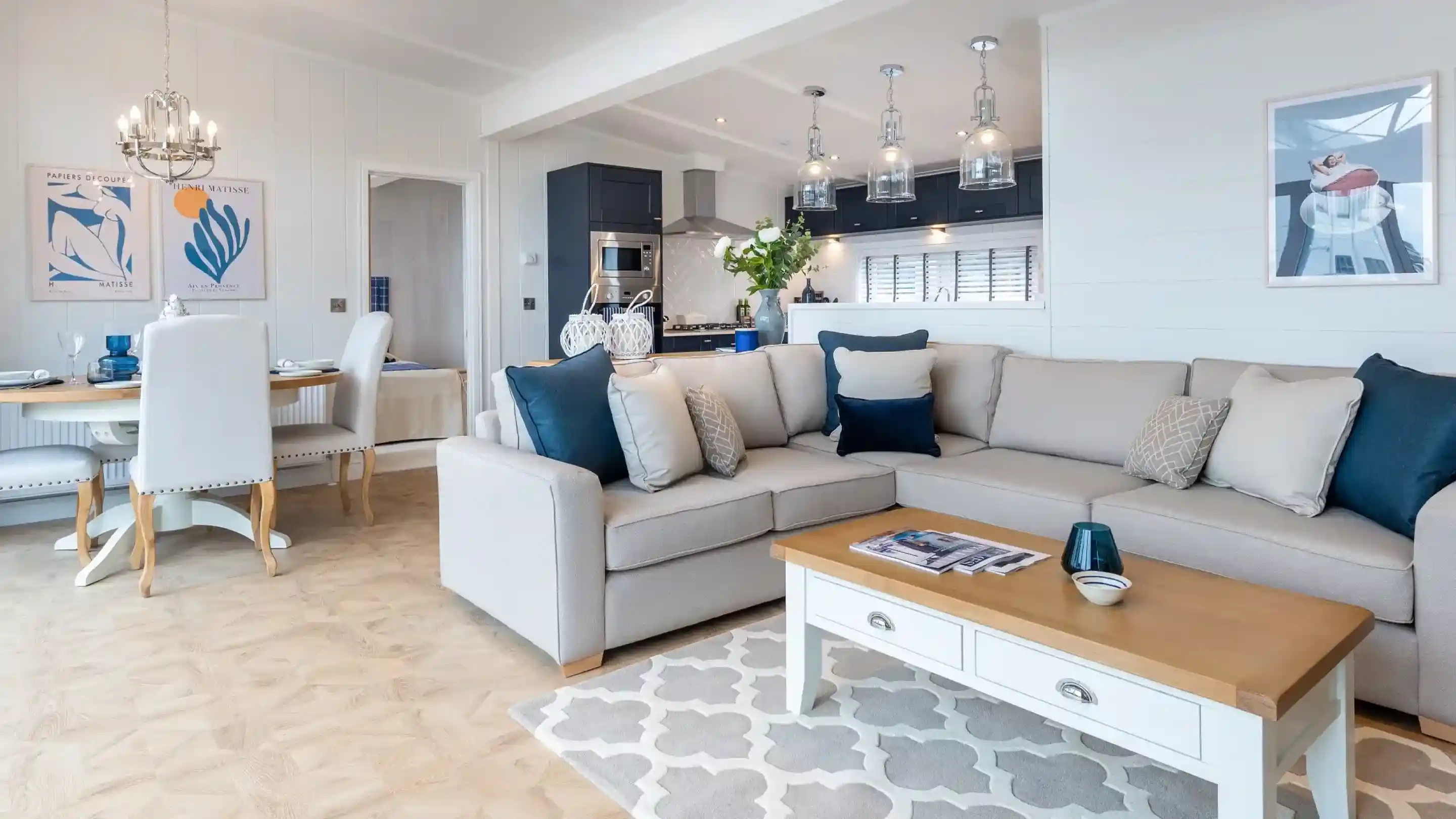 A stylish living room with a light grey sectional sofa adorned with decorative pillows. A wooden coffee table sits in front, topped with magazines. The open space features a dining area with a round table and elegant chairs. Modern pendant lights hang from the ceiling, and artwork decorates the walls.