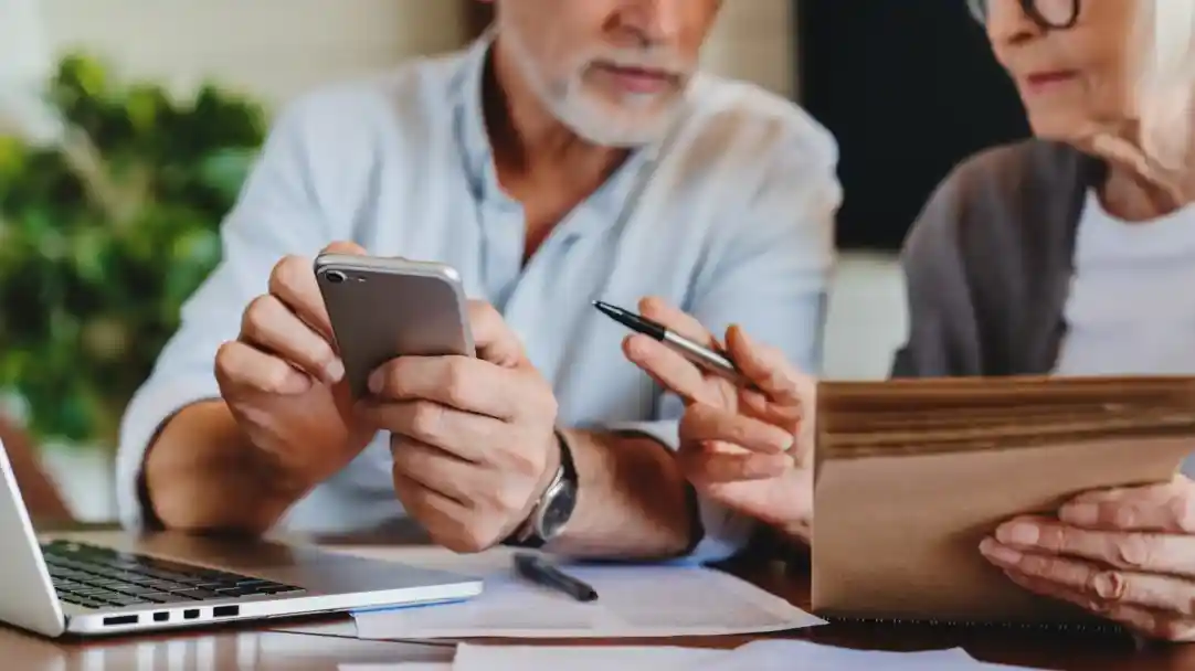 Two older adults engage in a discussion at a table, one holding a smartphone while the other points to notes in a notebook. A laptop sits open, along with scattered papers and a pen on the table.