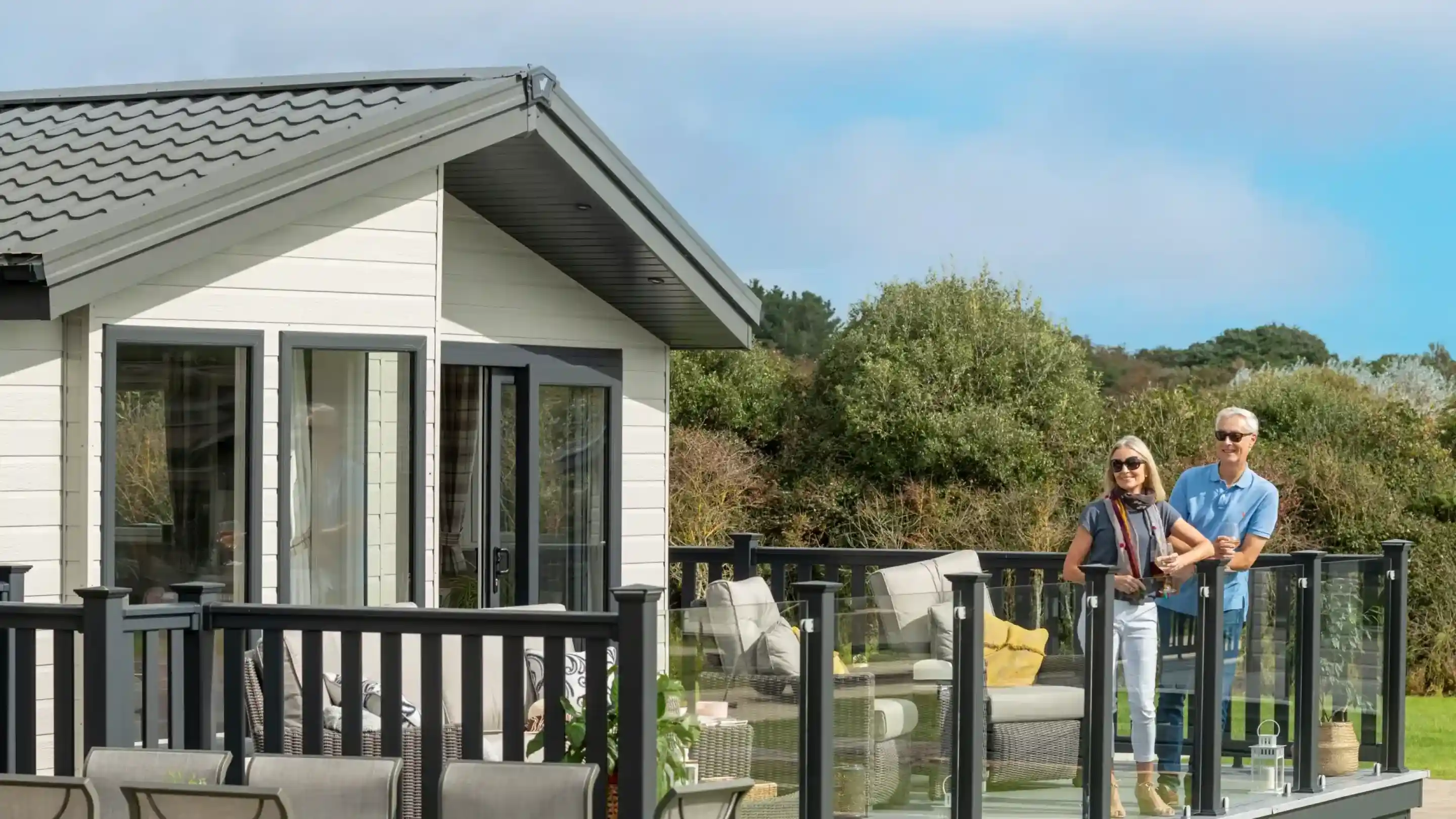 A couple stands on a balcony overlooking a green landscape. The woman wears sunglasses and a scarf, while the man is casually dressed. Behind them is a modern lodge holiday home with large windows and a sloped roof.