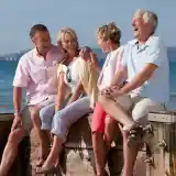 A group of four older adults joyfully sitting on a wooden dock by the beach, smiling and engaging with each other. The sun is shining and the ocean stretches out behind them.