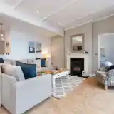 Bright and cozy living room featuring a light-colored sofa with blue and gray cushions, a wooden coffee table, a patterned rug, and a decorative fireplace. Soft lighting and neutral walls create a warm atmosphere, with a glimpse of a hallway and an armchair in the background.