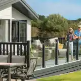 A couple stands on a modern deck with glass railings, overlooking a lush green landscape. The woman has long, blonde hair and is wearing a casual outfit, while the man has gray hair and is dressed in a light blue shirt. A stylish outdoor seating area is visible in the background.