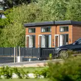 A modern wooden cabin with large windows sits amidst greenery, featuring a spacious deck and a black SUV parked nearby.