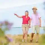 A couple walks hand in hand along a grassy path near the water, smiling and engaging in conversation. One person points to something in the distance, while the other looks on, both dressed casually for warm weather.
