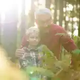 A senior man embraces a young boy in a sunlit forest, surrounded by ferns. The boy wears a checkered shirt and looks towards the camera, while the man smiles warmly beside him.