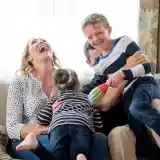 A family enjoys playtime at home, with a woman laughing joyfully while holding a young girl. A boy, smiling widely, is playfully engaged with a man in the background. Bright natural light streams through a window, creating a cheerful atmosphere.