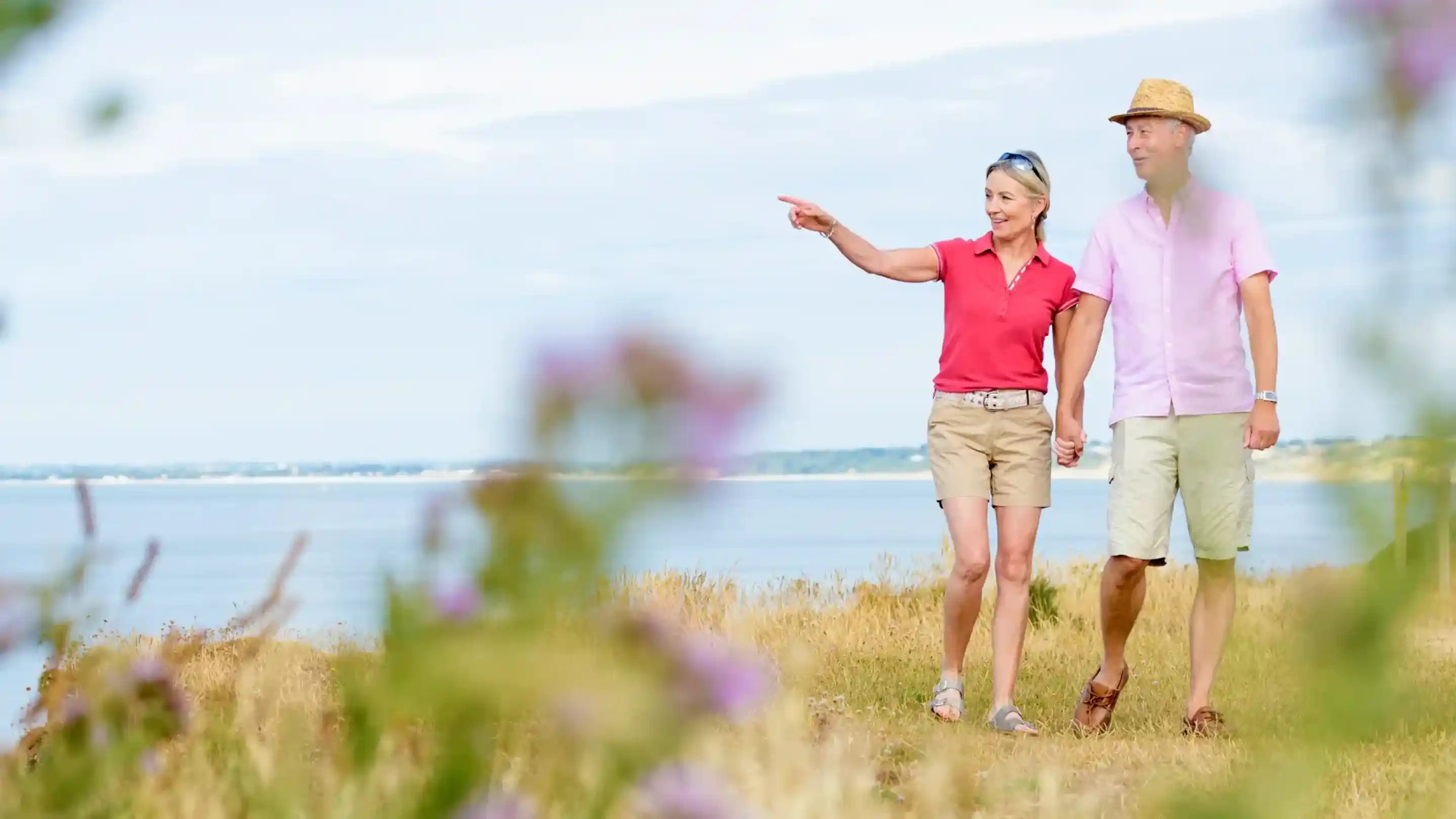 A couple strolls along a scenic coastline, hand in hand. The woman points towards something in the distance while wearing a pink shirt and shorts, complemented by a straw hat. The man, in a light pink short-sleeve shirt and beige shorts, walks beside her, both surrounded by blooming wildflowers.