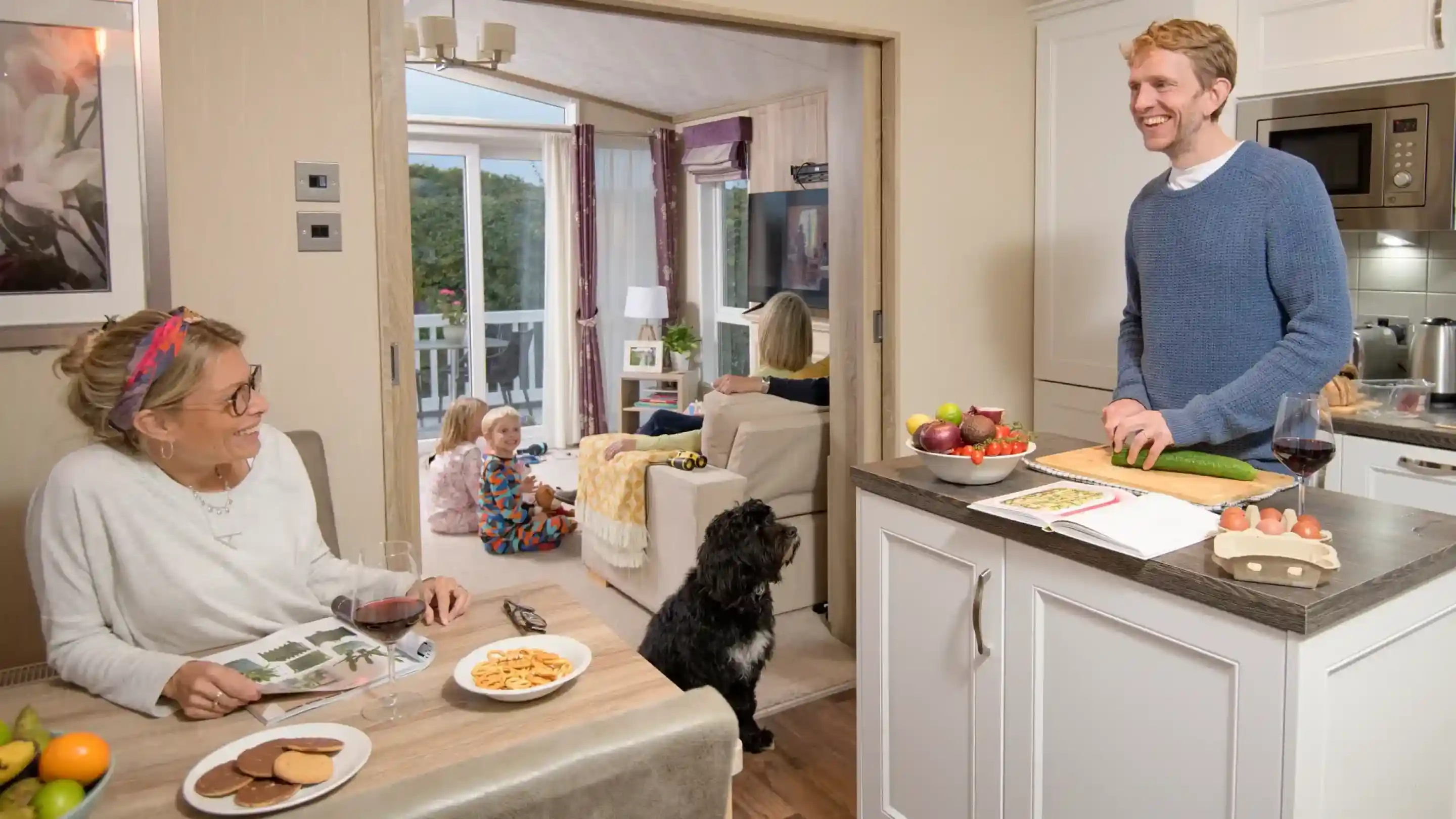 A man is smiling and standing in a modern kitchen, holding a cucumber. A woman sits at the kitchen island with a glass of wine and a plate of snacks, looking at him. In the background, a child plays and another person relaxes on a couch. A dog is lying on the floor nearby.