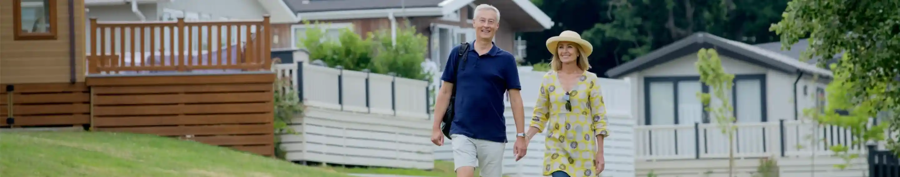 A couple walks hand in hand along a grassy path, surrounded by modern houses with wooden decks and greenery in the background. The woman wears a bright yellow dress and a wide-brimmed hat, while the man is casually dressed in a navy shirt and shorts.