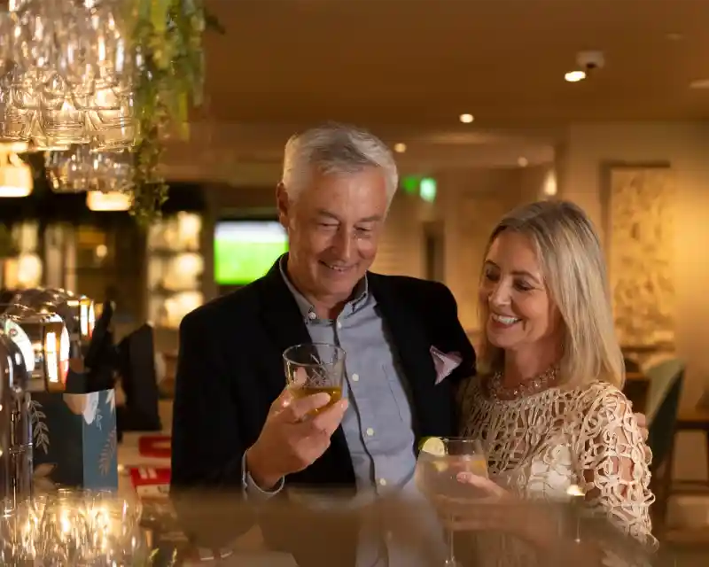A man and a woman share a joyful moment at a bar, each holding a drink. The man, with silver hair and a blazer, smiles as he looks at the woman, who wears a light-coloured lace top and has long, blonde hair. The background features softly lit décor and glassware.