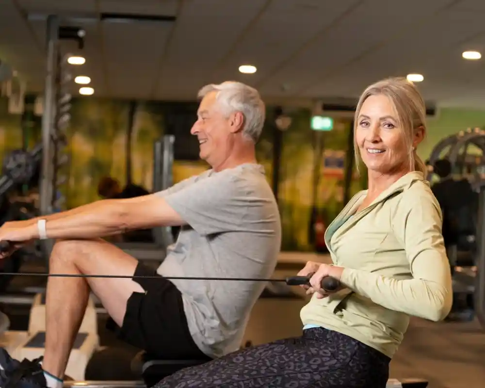 A man and a woman engage in a rowing exercise at a gym. The man is in the background, smiling and pulling the rowing handle, while the woman is in the foreground, looking at the camera with a confident expression. Both are dressed in athletic clothing.