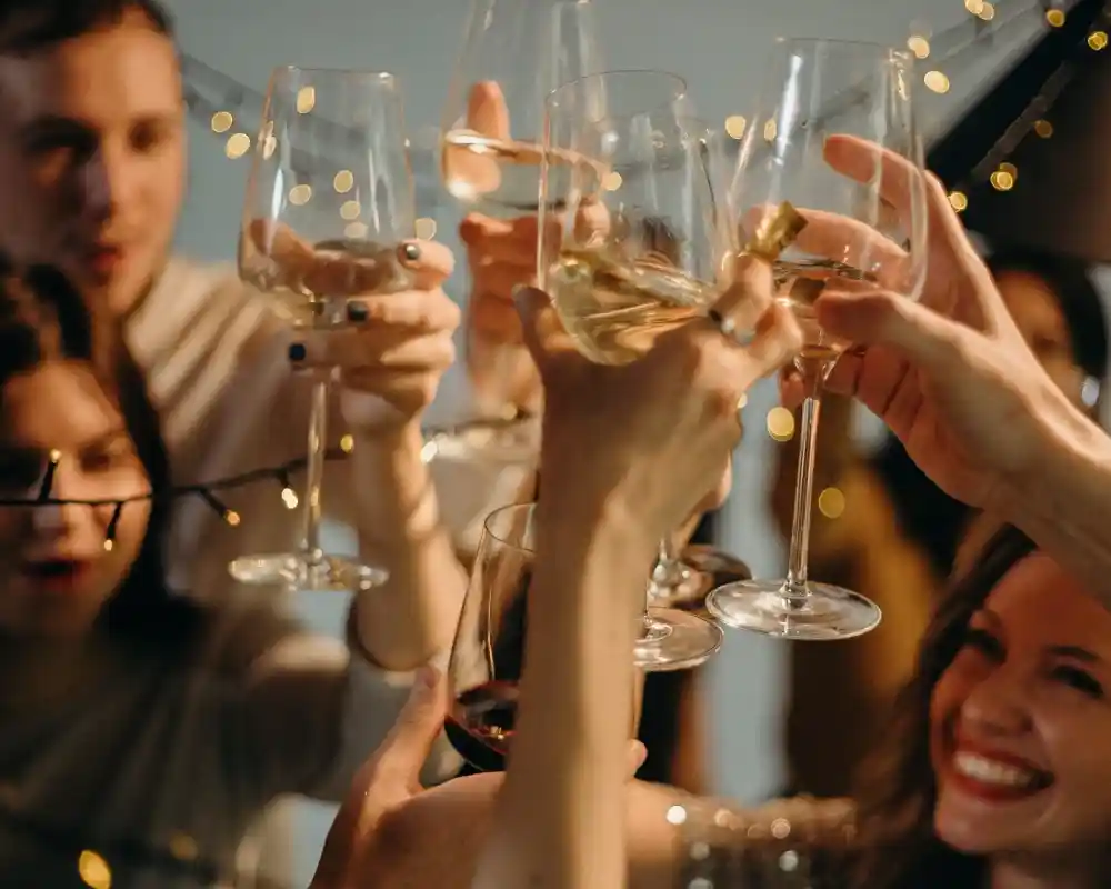 A group of people celebrating, raising glasses filled with different drinks in a toast. Soft string lights are visible in the background, creating a festive atmosphere.
