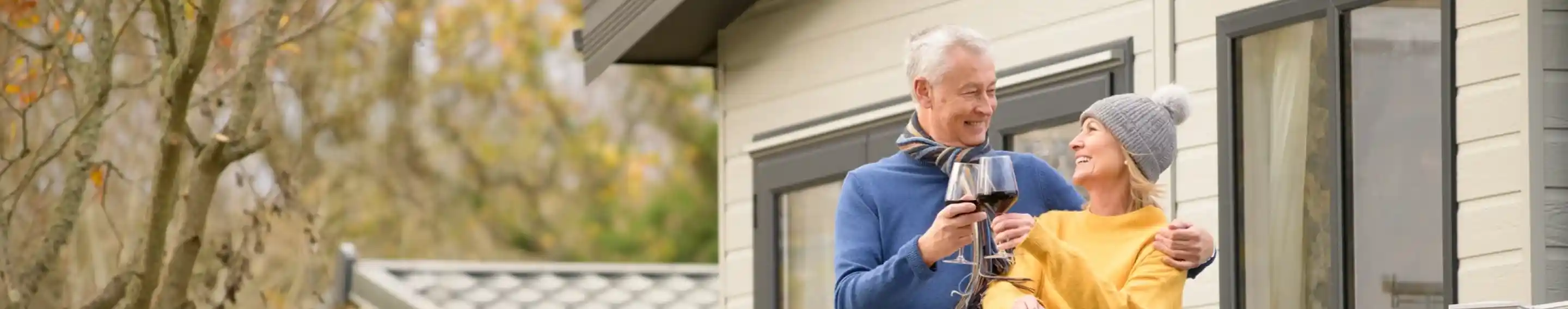 A happy couple stands on a balcony, enjoying drinks together. The man is wearing a blue sweater and holding a glass, while the woman is in a yellow sweater and a gray beanie. They share a joyful moment with a house and autumn trees in the background.