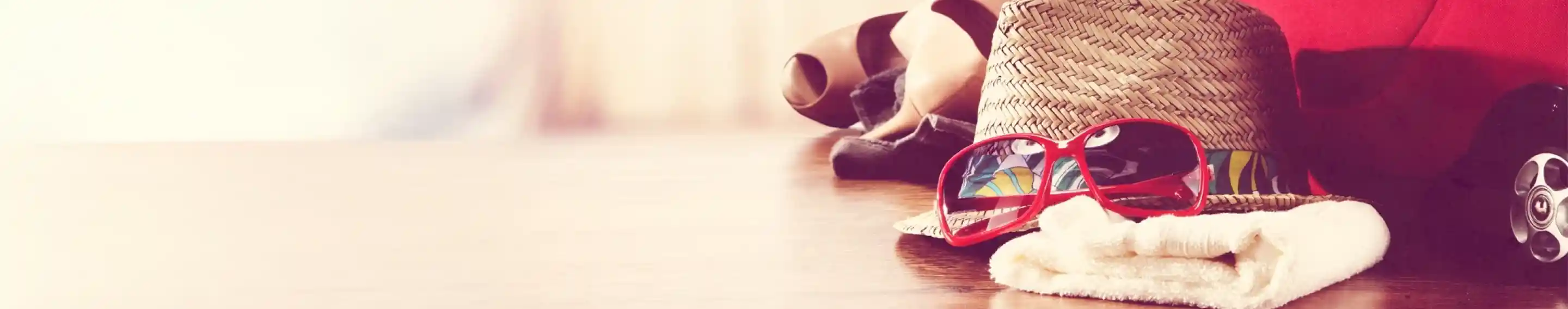 A straw hat and colorful sunglasses sit on a wooden table, surrounded by beach towels and a red cooler. The background is softly blurred, suggesting a relaxed summer atmosphere.