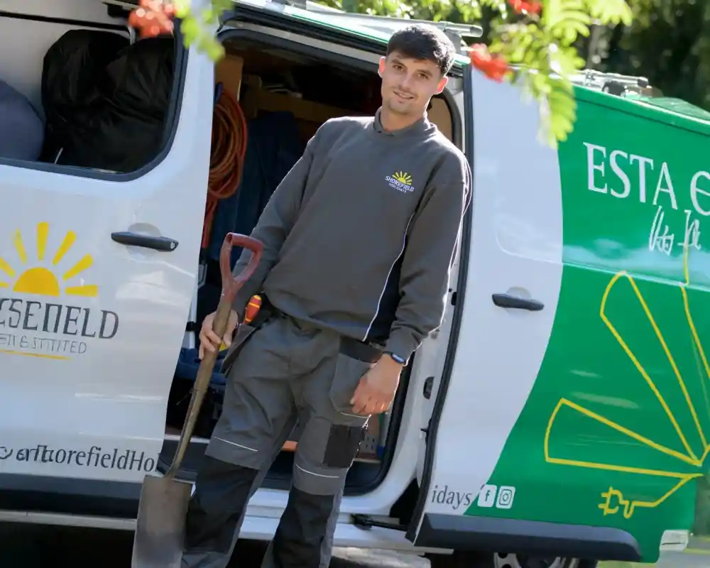A young man in a gray uniform stands next to a brightly colored van. He holds a shovel and smiles at the camera. The van features a sun logo and text in Spanish. Tools and equipment are visible inside the van.