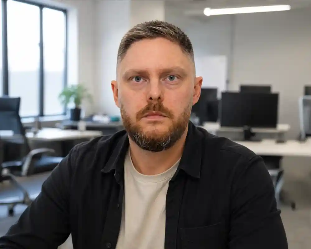 A serious man with short, light brown hair and a beard sits at a table in an office environment. He wears a black shirt over a light-colored t-shirt. In the background, there are empty desks with computers and a plant near a window.
