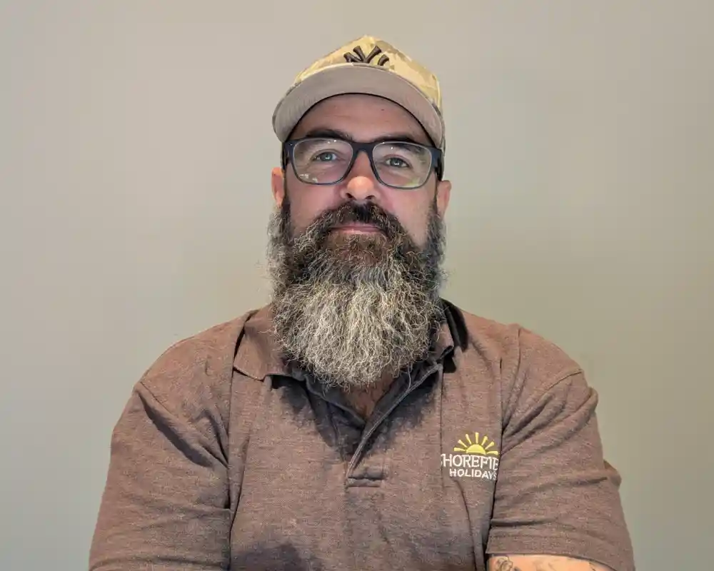 A man with a thick gray beard and glasses wears a cap and a brown polo shirt with a logo. He has his arms crossed and is sitting against a light gray wall.